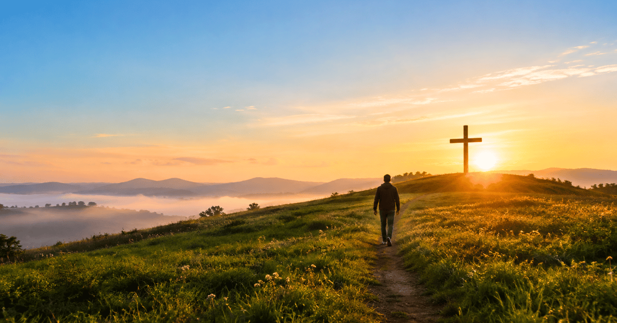 Pessoa caminhando por uma trilha em um campo verde em direção a uma cruz de madeira no alto de uma colina ao nascer do sol.